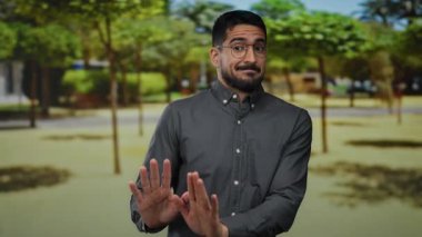 Parkta duran genç adam, elleri havada, gözlüklü ve gri gömlekli, güneşli bir gökyüzünün altında yeşil ağaçlarla çevrili, düşünceli görünüyor..