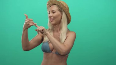 Yeşil stüdyoda telefon hareketlerini gösteren eli kulağında hasır şapka takan sarışın bir kadın..