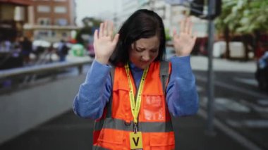 Güvenlik yeleği giymiş esmer saçlı bir kadın kalabalık bir caddede elleri havada, açık bir şehir ortamında reddedilerek duruyor..