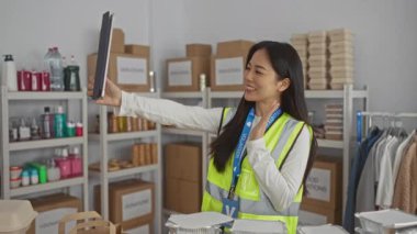 Yansıtıcı yelek giyen genç Çinli bir kadın, organize bağışlar ve malzemelerle çevrili bir hayır kurumunun gönüllü merkezinde video konferansı düzenliyor..