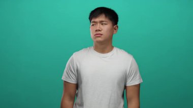 Gündelik kıyafetli genç Çinli adam canlı yeşil bir arka plana karşı şaşkın duruyor, dalgın ve şaşkın görünüyor..