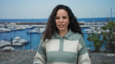 Genç İspanyol kadın, arka planda teknelerle bir sahil limanının yakınında neşeli bir şekilde gülümsüyor. Açık hava ortamında rahat ve keyifli bir şekilde çizgili bir süveter giyiyor..