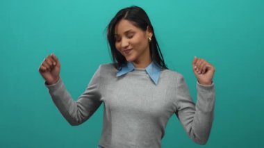 İzole edilmiş yeşil bir duvara karşı neşeyle dans eden, uzun siyah saçlı, gri bir kazak giyen, stüdyoda mutluluk ve pozitiflik saçan bir kadın..