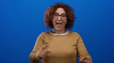 Orta yaşlı İspanyol bir kadın heyecanlı bir şekilde her iki eliyle canlı bir mavi arka plana karşı hareketli, bir stüdyo ortamında pozitiflik ve enerji iletiyor.