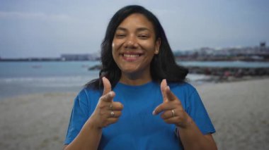 Mavi gömlekli bir kadın güneşli bir kumsalda işaret parmağıyla kamerayı işaret ediyor..