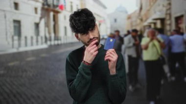 Şehir caddesinde kredi kartı tutan genç adam mali karar üzerinde kafa yoruyor. İnsanları ve şehir planlarını göz önünde bulundurarak açık hava ortamını ve düşünceli ifadeyi öneriyor.