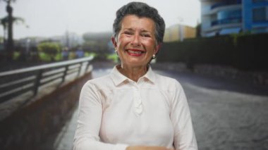 Deniz kenarında gezinirken zarif bir şekilde gülümseyen gri saçlı yaşlı kadın, berrak bir gökyüzünün altında kumsalın dingin güzelliğiyle çevrili..