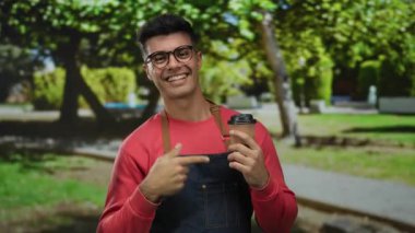 Kırmızı kazaklı genç adam yeşil bir parkta kahve fincanı tutuyor güneşli bir günde arka planda ağaçlar ve çimlerle gülümsüyor..