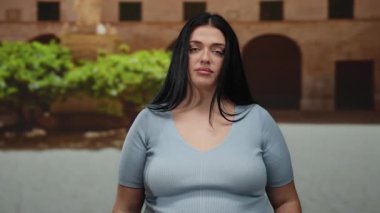 Şehir sokaklarında elini başını gösteren parmaklarıyla el kol hareketi yapan, hayal kırıklığını ya da tefekküre dalmayı ifade eden, artı boy ve kıvrımlı vücut, günlük mavi bluzlar giyen bir kadın..