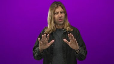 Sarışın, uzun saçlı bir adam ciddi bir ifadeyle mor stüdyo duvarına el kaldırıyor..