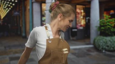 Önlüklü genç sarışın kadın işlek bir şehir caddesinde neşeli bir şekilde gülümsüyor. Parlak vitrinler ortamı hareketli ve davetkar kılıyor..