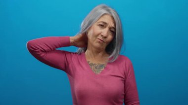 Parlak mavi arka planda, eli boynundaydı. Gri saçları ve pembe süveteri vardı. Düşünceli bir yüz ifadesi vardı..