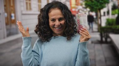 Şehrin dışında bir kadın gülümseyerek saçlarını tarıyor ve elinde yeni Zelanda dolarları, arka planda canlı bir şehir manzarası, şehir manzarasından keyif alıyor.