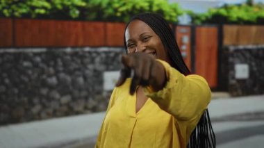 Sarı tişörtlü kadın, güneşli bir günde şehir sokağında taş duvar ve bitki örtüsüyle kameraya bakıp gülümsüyor..