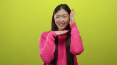 Sarı arka planda pembe süveter giyen, Asyalı yüz hatları ve izole stüdyo ortamında uzun siyah saçlı neşeli ve pozitif ifade sergileyen bir kadın..