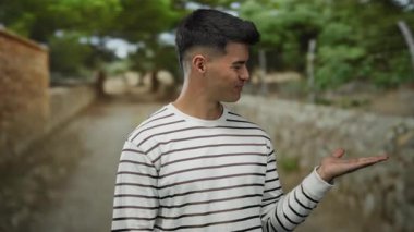 Çizgili tişörtlü ve elini açık bir parkta dikilen genç bir adam. Arka planda taştan bir yol ve yemyeşil bir zemin var. Neşeli görünüyor..