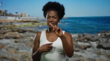 Mavi gökyüzünün altındaki kayalık plajda el kol hareketi yapan bir kadın, doğal deniz manzarası sergiliyor, ifadesini ve sahil ortamında dış görünüşünü vurguluyor..