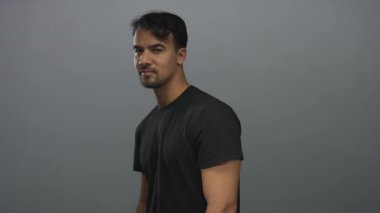 İzole edilmiş, gri arka planda, siyah gömlekli, arkadaş canlısı tavırları ve olumlu tavırları olan İspanyol asıllı genç adam güvenle gülümsüyor.