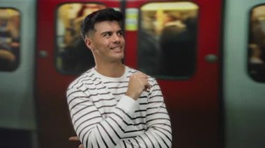 Tren istasyonunda gülümseyen genç bir adam, çizgili bir gömlek giyiyor, bulanık bir arka plan yolcuları ve hareket halindeki bir tren, şehir seyahatinin ve tarzının özünü yakalıyor..