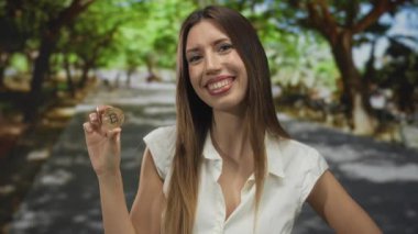 Genç kadın, güneş ışığıyla kaplı bir caddede ince parmakları arasında parlak bitcoin tutuyor; dijital zenginlik iyimserliği.