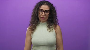 Kahverengi kıvırcık saçlı ve gözlüklü genç bir kadın mor stüdyo ortamında el çırpıyor..