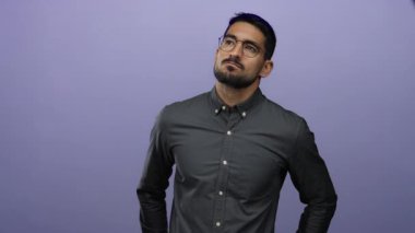 Gözlüklü genç adam yumuşak mor bir arka plan üzerinde düşünceli bir şekilde kafa yoruyor, düşünceli ve odaklanmış bir atmosfer yaratıyor..