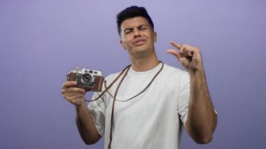 Beyaz gömlekli İspanyol adam, izole edilmiş mor arka plana karşı elinde klasik kamera tutarken parmaklarıyla küçük bir jest yapıyor..