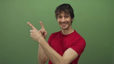 Kırmızı tişörtlü, geniş gülümsemeli bir adam her iki işaret parmağını stüdyoya işaret ediyor; coşku ve pozitif enerji..