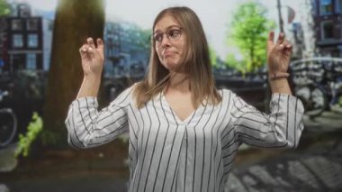 Genç sarışın kadın gün ışığında iki eliyle bir sokak kanalında tırnak işareti yapıyor, çizgili üst ve yuvarlak gözlükler takıyor; şakacı ironi..