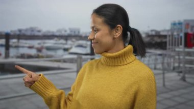 Sarı süveterli bir kadın limana yanaşan uzak tekneleri işaret ederek bulutlu deniz manzaralı sakin bir açık hava yaratıyor ve görünürde gemiler var..