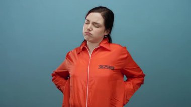 Turuncu hapishane tulumu giyen kadın mavi arka planda duruyor. Yüzünde acı dolu bir ifade ve sırtında ellerle..