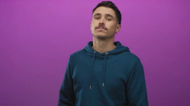 Deniz mavisi kapüşonlu genç İspanyol adam canlı mor arka plana karşı duruyor, izole bir ortamda günlük tarz ve güven yayıyor..