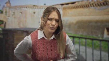 Midesi ağrıyan genç bir kadın antik Roma harabelerinin önünde duruyor, açık şehir ortamında rahatsızlık gösteriyor, pembe bir kazak ve beyaz gömlek giyiyor..