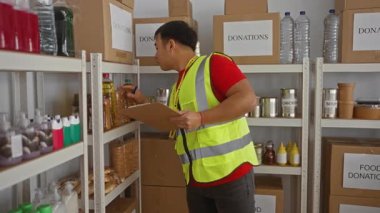 Bağış merkezinde çalışan genç bir adam yansıtıcı yelek giyiyor ve düzgün düzenlenmiş yiyecek eşyaları ve kutular arasında bir pano tutuyor..