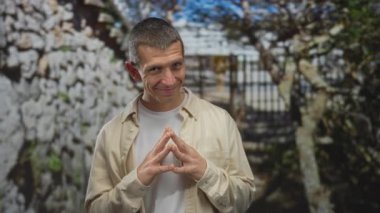 Güneşli bir günde taş duvar ve ağaçlarla dolu açık havada gülümseyen bir adam, günlük gömlek, sokak arkası, rahat duruş, doğanın keyfi, elleri bağlı, huzurlu bir ifade, huzurlu bir ortam.