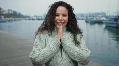 Deniz kenarında sıcacık gülümseyen bir kadın, manzaralı bir limanda rahat bir süveter giyiyor, kayıklar nazikçe yüzüyor, sakin su ve açık hava sükunetiyle çevrili..
