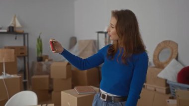 Yeni eve video alan bir kadın. Oturma odasındaki kutuların arasında günlük kıyafetleri ve telefonları taşıyor..