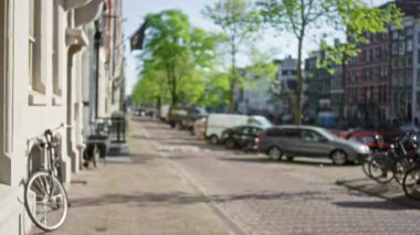 Amsterdam 'da güneşli bir caddenin bulanık manzarası park halindeki bisiklet ve arabalarla Hollanda' daki bir kanal boyunca uzanan ağaçların ve tarihi binaların önünde..