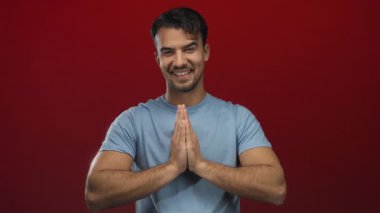 Genç İspanyol adam, elleri namaste jesti ile gülümsüyor mavi gömlekli, soyutlanmış kırmızı arka plana karşı minnettarlığını ve huzurunu ifade ediyor.