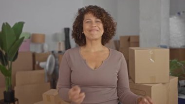 Kolları bağlı bir şekilde gülümseyen ve yeni bir binanın içinde karton kutuları taşıyan bir kadın..