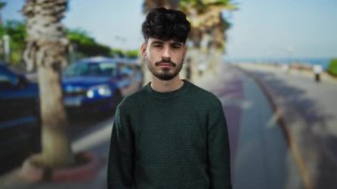 Arka planda palmiye ağaçları olan kıvırcık saçlı yakışıklı genç adam rahat ama düşünceli bir tavır sergiliyor..