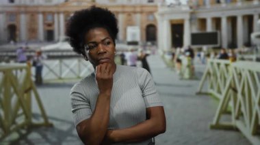 Kadın titiz bir şekilde Vatikan 'ın dış mekanında dikiliyor. İkonik mimarisi görünür ve arka planda insanlar bir anlık tefekkür sergiliyor..