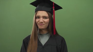 Stüdyoda burnu buruşmuş genç bir kız yeşil bir fon karşısında mezuniyet kepi ve cübbe giyerken yüzünü buruşturuyor..