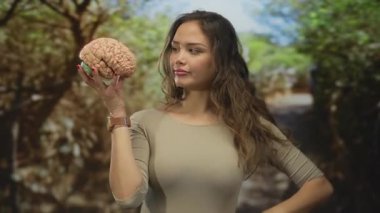 Parkta elinde saatli bir beyin mankeni olan bir kadın, İspanyol kökenli, bej giyen, açık hava ortamında yemyeşil alanlar arasında düşünceli bir şekilde gezinen..