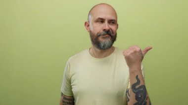 Kel sakallı, dövmeli, başparmağı sarı duvara dayalı, izole bir arka planı olan bir adam.