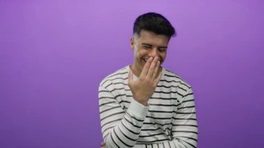 Genç adam el üstünde gülüyor, çizgili süveter giyiyor, canlı mor bir arka plana karşı, bir stüdyo ortamında neşeli bir ifade sergiliyor.