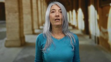 Eski bir üniversitenin açık hava koridorunda elleri dramatik bir şekilde kır saçlı, turkuaz süveter giyen son sınıf öğrencisi bir kadın..