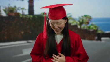 Kırmızı mezuniyet elbisesi giymiş Asyalı genç bir kadın şehir arka planındaki şehir sokağında gülümsüyor.