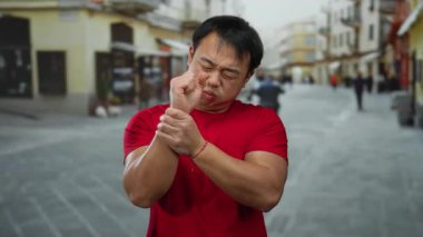 Kırmızı gömlekli genç adam şehir caddesinde bileğini tutarak acı içinde duruyor. Arka planda bulanık binalar ve insanların olduğu şehir ortamını gösteriyor..