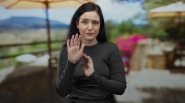 Esmer bir kadın, güneşli bir restoranın terasında, arka planda dağlar olan manzaralı bir caddeye bakarak anlamlı bir şekilde el kol hareketi yapıyor, duygularını iletiyor..